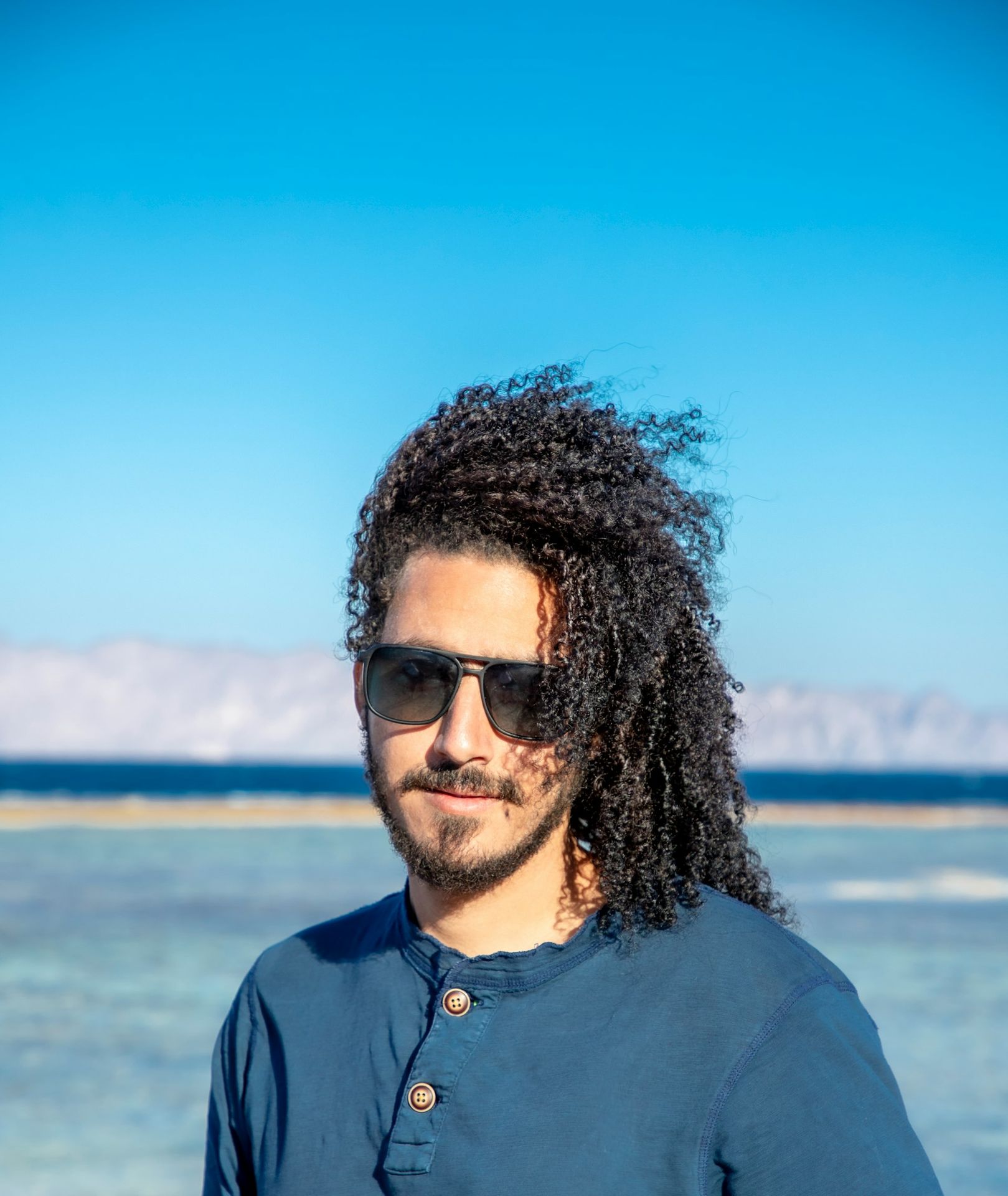 a man with long hair and sunglasses standing in front of a body of water